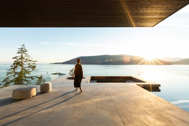 A minimalist waterfront terrace or pool deck viewed from beneath a broad timber-lined roof overhang. The open concrete platform overlooks calm water and a mountainous or hilly shoreline, with a person walking toward the edge, built-in low seating, and a rectangular recessed pool or water feature. The scene is strongly connected to the landscape, with warm low-angle sunlight and a serene contemporary atmosphere.