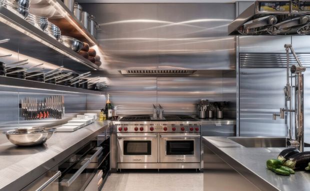 Professional commercial-style kitchen with extensive stainless-steel wall cladding, open metal shelving, restaurant range and ovens, deep sink, commercial ventilation hood, and organised cooking equipment. The space has a highly functional, industrial fit-out with cool metallic surfaces and bright task lighting.