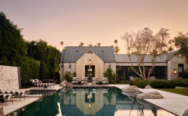 A refined single-storey courtyard-style residence viewed from across a swimming pool at dusk. The central gabled pavilion has pale vertical timber cladding and a dark standing-seam metal roof, flanked by low wings with light masonry walls and matching metal roofing. Large glazed openings connect to furnished outdoor terraces, with a landscaped garden, boulders, and pool reflections in the foreground.