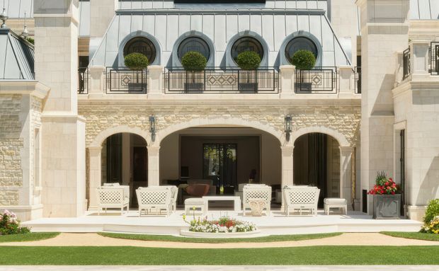 A grand, symmetrical stone-clad courtyard facade with a covered outdoor terrace. The architecture features arched openings, ornate dark metal balustrades, round-top dormer-like windows, pale masonry, and a formal landscaped lawn with clipped topiary and flower planters. White outdoor lounge chairs and a central seating arrangement occupy the terrace.