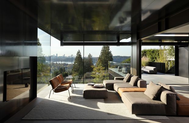 A refined contemporary living room with a large sectional sofa, sculptural lounge chair, and low ottoman arranged around a dark built-in fireplace wall. Floor-to-ceiling glazing opens to expansive distant views, while dark ceiling and wall surfaces contrast with pale upholstered furniture and a light rug. An adjacent outdoor terrace or covered outdoor area is visible through the opening on the right.