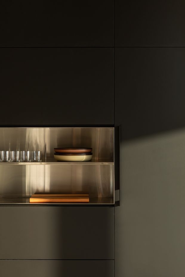 A close-up of sleek, dark built-in cabinetry with an illuminated open display niche. The niche has reflective metallic lining and contains neatly arranged glassware, stacked bowls, and a wooden serving board. Strong directional sunlight creates warm reflections and deep shadows, giving the minimalist joinery a refined, moody character.
