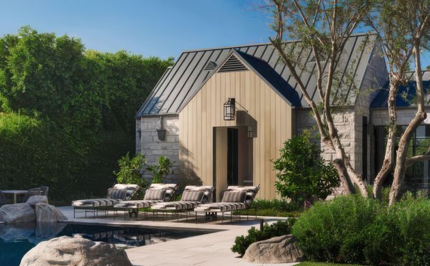 Exterior view of a refined poolside pavilion or residence with a steep gabled metal roof, vertical light timber cladding, pale stone walls, dark-framed glazing, and a landscaped terrace with loungers beside the swimming pool. Mature planting and rock formations surround the outdoor area.