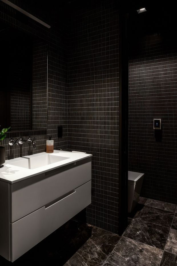 A dark, dramatically lit contemporary bathroom featuring a floating grey vanity with an integrated white basin and wall-mounted chrome tapware. The walls are covered in narrow glossy black grid-pattern tiles, while the floor has dark veined stone-look tiles. A partially screened toilet area is visible to the right, with a mirror and minimal black fixtures completing the moody scheme.
