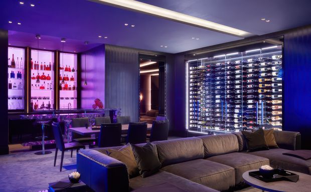 A luxurious, moody entertainment lounge with deep purple and blue ambient lighting. The room features a large sectional sofa in the foreground, a dining or games table with dark upholstered chairs, illuminated glass-front wine storage displaying numerous bottles, and backlit liquor shelving with a bar counter. Recessed ceiling lighting and glossy dark finishes create a dramatic high-end atmosphere.