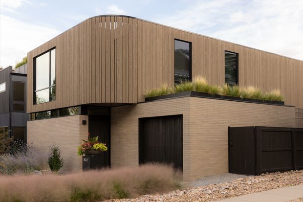 Contemporary two-storey residence with a sculptural curved upper volume clad in vertical timber battens, projecting over a lower level of pale brickwork. The facade features dark-framed windows, a recessed entry, an integrated garage, black fencing and a planted roof terrace. Ornamental grasses and a gravel forecourt create a refined, low-maintenance landscape.