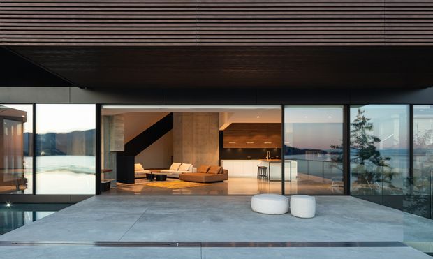 Contemporary open-plan residence viewed from a broad concrete terrace, with extensive floor-to-ceiling glazing opening onto a living area and kitchen. The interior features a low modular sofa, timber cabinetry, warm wood flooring and ceiling lining, while a dark sculptural stair or fireplace wall creates contrast. Water and distant landscape are visible beyond the glazing.