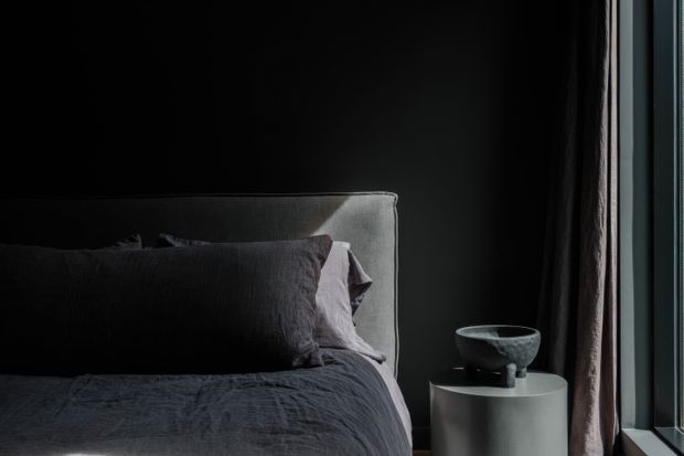 Moody close-up of a contemporary bedroom corner, featuring a dark upholstered bed with layered charcoal bedding and pillows, a black wall, and a small white cylindrical bedside table holding a dark sculptural vessel. Floor-to-ceiling dark curtains frame a narrow window at right, contributing a strong vertical light contrast.