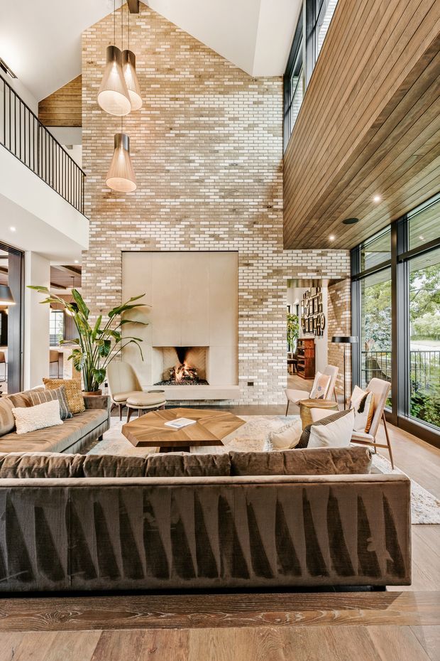 A spacious double-height living room with a large white and tan brick fireplace wall as the focal point. The room features expansive black-framed glazing, a warm timber-clad wall and ceiling, pendant lights, exposed timber flooring, a large sectional sofa, lounge chairs, a timber coffee table, and a mezzanine-level black metal balustrade. The overall design blends contemporary architecture with warm natural materials and a relaxed, refined interior.