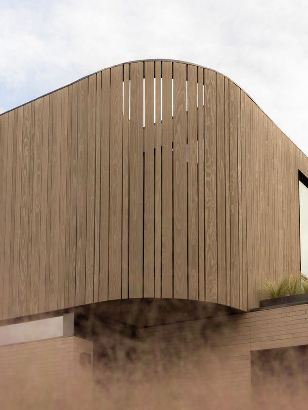 Close-up exterior view of a contemporary building facade featuring a dramatic curved, arched roofline or upper wall clad in vertical timber slats. The slatted timber screen creates narrow openings and conceals parts of the structure, while pale brickwork is visible below and at the side. Minimal planting appears along the lower edge, with an overcast sky and slight foreground haze.