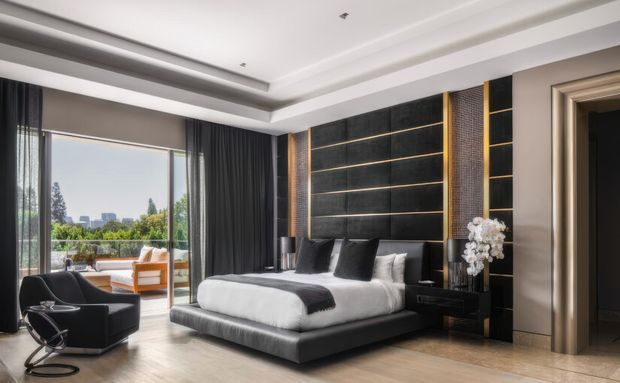 A sophisticated contemporary bedroom with a large upholstered platform bed, black and white bedding, and a dramatic dark feature wall with horizontal shelving and warm metallic trim. Floor-to-ceiling glazing opens to a balcony or terrace, bringing in daylight and views of greenery and distant buildings. The room also includes dark curtains, a sculptural lounge chair, bedside furniture, decorative lighting, and a refined layered ceiling.