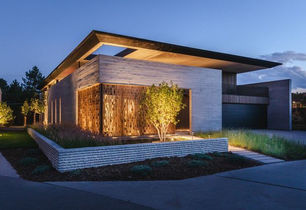 Twilight exterior view of a contemporary single-storey residence with a dramatic cantilevered flat roof, pale horizontal cladding, warm timber screening at the entry, deep roof overhangs, and an integrated garage. The home is set within a landscaped front garden with raised textured retaining planters and a paved path.