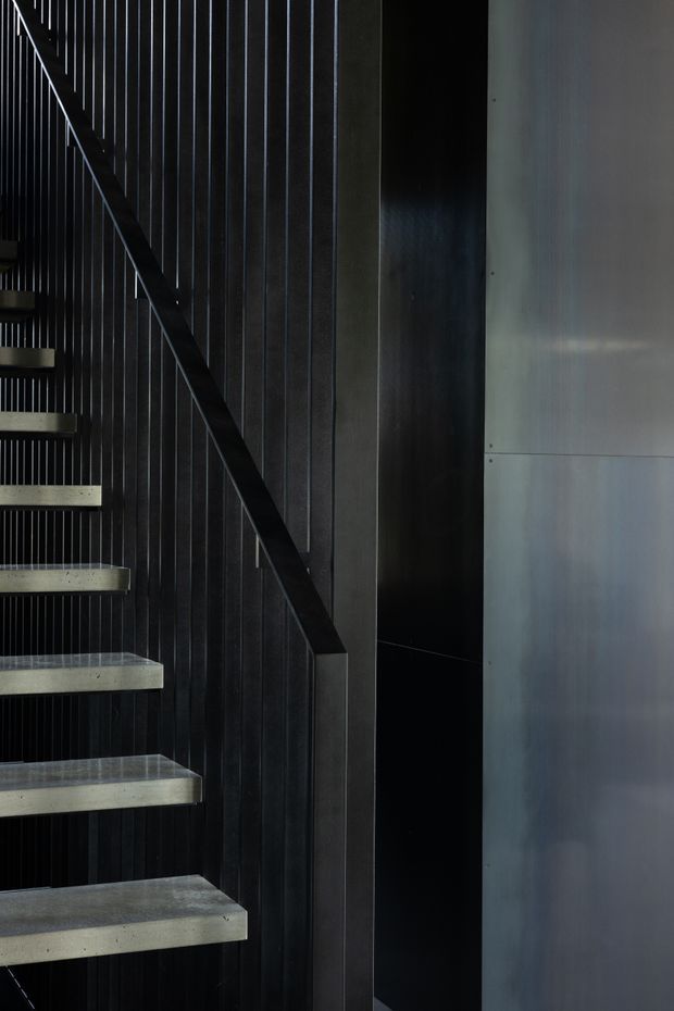 Close-up architectural interior view of a modern floating staircase. Pale concrete treads project from a dark, vertically ribbed wall, with a slim dark handrail and black recessed void beside a smooth grey wall panel. The composition is stark, refined, and highly geometric, with dramatic low-key lighting.