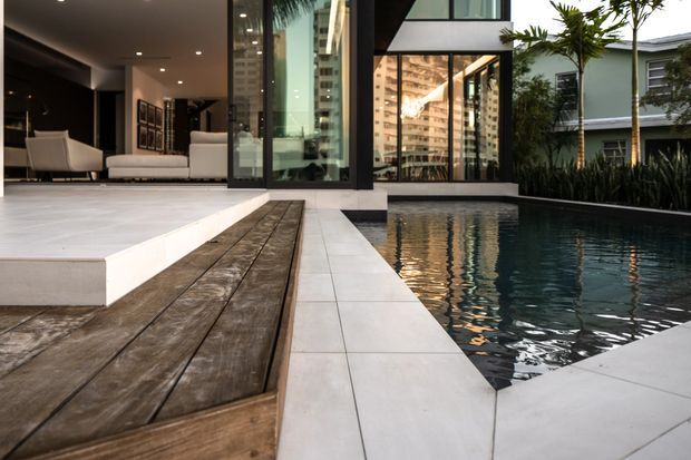 Contemporary residential indoor-outdoor living area opening onto a narrow reflecting or swimming pool. The scene features large floor-to-ceiling glass walls, a white tiled terrace, a raised timber deck walkway, dark interior finishes, and lush tropical planting beside the pool. A living room is visible through the glazing, while the pool terrace dominates the composition.