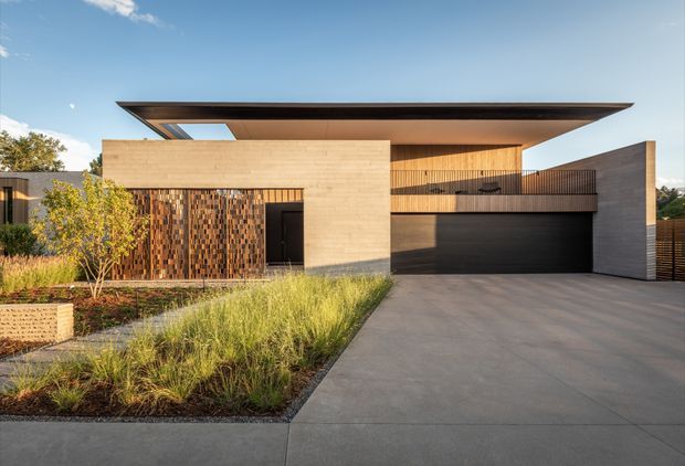 Front exterior view of a contemporary multi-level residence with a dramatic thin cantilevered flat roof, pale masonry walls, warm timber-lined upper level, dark metal garage door, and a textured vertical brick feature wall. A broad concrete driveway and landscaped native-grass garden lead to the recessed entry and double garage.