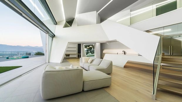 A bright, highly contemporary open-plan living space with sculptural angular white walls and ceilings, floor-to-ceiling glazing opening to an outdoor terrace and distant landscape, pale timber flooring, integrated shelving, and a floating staircase with glass balustrade. A large curved modular sofa occupies the centre, while linear recessed lighting emphasizes the geometric architecture and double-height volume.