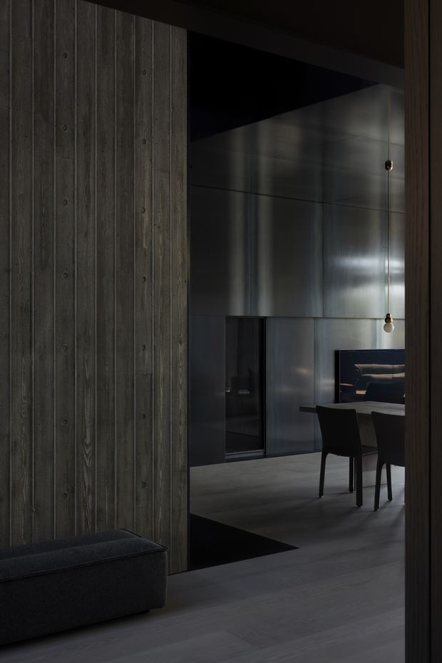 A moody, dark-toned interior view framed through a doorway, featuring vertical weathered timber wall panelling, dark flooring, and a partially visible dining area with a dark table and chairs. Metallic wall cladding, pendant lights, and a glimpse of a lounge or seating area add a refined industrial character.