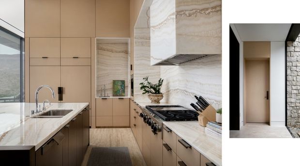 A refined galley kitchen with warm beige built-in cabinetry, pale veined stone benchtops and matching full-height splashback. The kitchen includes a central island with an undermount sink and curved chrome tap, integrated storage, a large cooktop, and a sculptural stone-clad rangehood. A narrow inset image at right shows a minimalist interior doorway beside a natural stone wall. Large glazing at left provides views toward a mountainous landscape.