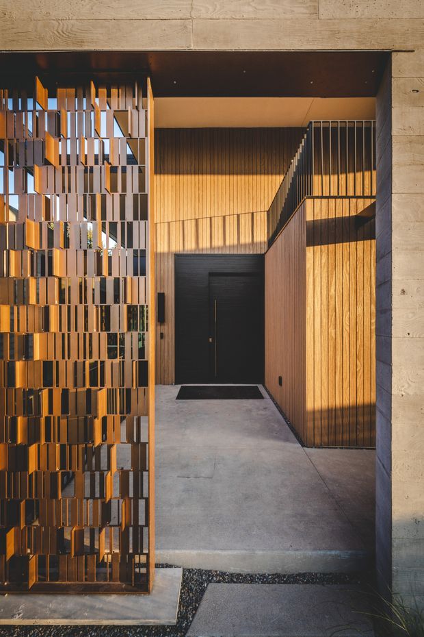 Architectural photograph of a refined residential entryway framed by a concrete portal. A perforated metal screen gate with staggered rectangular elements sits to the left, while warm vertical timber wall cladding and a tall black entry door create a layered entrance. Concrete paving, dark gravel strips, and a black metal stair or balcony railing complete the modern composition, with strong low-angle sunlight creating warm shadows.