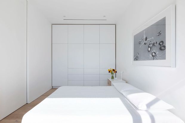 Bright minimalist bedroom with a low-profile white bed, crisp white bedding, pale timber flooring, and full-height white built-in wardrobes. A large monochrome artwork hangs on the wall, while concealed linear lighting and streamlined cabinetry reinforce the clean, pared-back design.