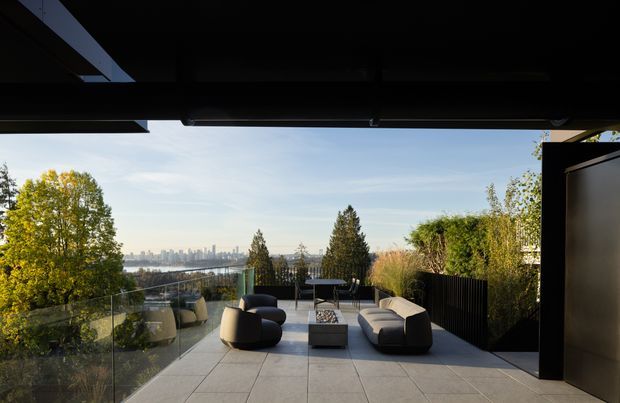 A contemporary elevated outdoor terrace viewed from beneath a dark roof overhang, furnished with curved lounge seating and a low coffee table. Large-format pale paving and clear glass balustrades define the terrace, with dense planting and a distant urban skyline visible beyond.