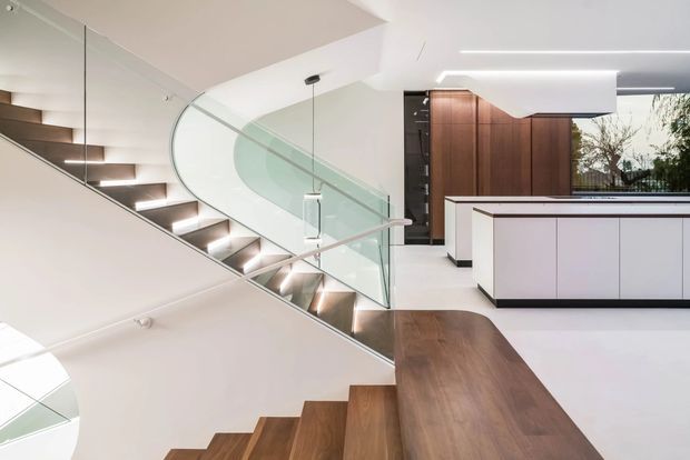 A contemporary open-plan interior focused on a sculptural timber staircase with illuminated treads and curved clear-glass balustrading. The stair connects multiple levels beside a minimalist white kitchen or built-in joinery zone, with warm dark timber wall cabinetry and a large timber surface in the foreground. Linear recessed lighting and white surfaces create a bright, refined setting.