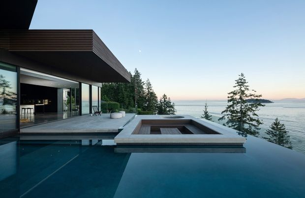 Contemporary coastal residence viewed from an outdoor pool terrace, with broad floor-to-ceiling glazing opening onto a minimalist living or entertaining area. A dark-edged infinity pool dominates the foreground, incorporating a raised rectangular sunken feature, while a cantilevered flat roof with horizontal timber-like soffit screening shelters the terrace. Coniferous trees and a broad body of water are visible beyond the property.