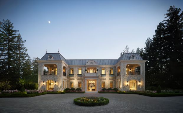 Symmetrical grand mansion facade viewed from a circular landscaped driveway at dusk. The building features pale stone masonry, a central entrance pavilion, two projecting wings with open arched loggias, tall multi-pane windows, ornate classical detailing, and steep metal-clad hipped roofs. Formal gardens, mature trees, exterior lighting, and a small central planted roundabout are visible.