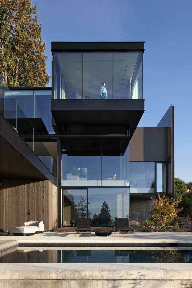 A dramatic contemporary multi-level residence viewed from the pool terrace. The home features strong cantilevered volumes, extensive floor-to-ceiling glazing, dark metal cladding and trim, timber wall cladding, and crisp geometric forms. Outdoor lounge furniture sits beside a long reflecting or swimming pool, with landscaped planting around the building.