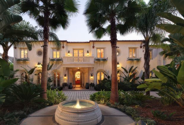 A symmetrical Mediterranean-style mansion facade viewed through a lush tropical garden. The pale stucco two-storey building has arched doorways and windows, decorative balconies, circular wall details, warm exterior lighting, and a terracotta-tiled roof edge. A round tiered fountain anchors the foreground courtyard, framed by tall palms and dense tropical planting.