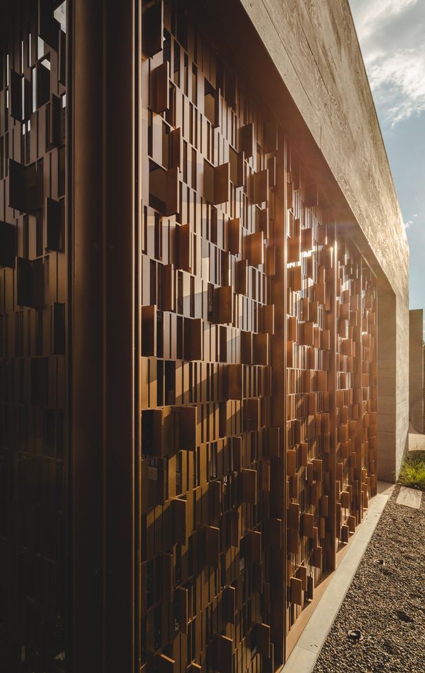 Close-up exterior architectural view of a contemporary building facade featuring a richly textured bronze-brown metal screen composed of many projecting vertical and rectangular elements. A large pale textured wall plane forms the upper edge, while a narrow gravel strip and concrete paving run alongside the facade. Strong low-angle sunlight creates dramatic highlights and shadows across the screen.