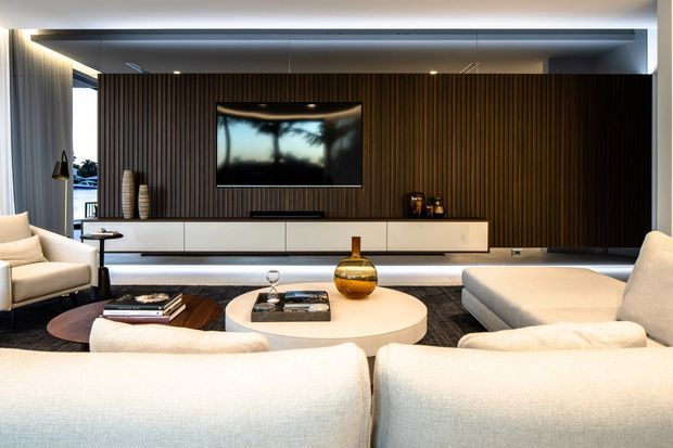 A refined contemporary living room viewed from behind a large cream sectional sofa. The room features a dark vertical timber-slat media wall with a wall-mounted television, a long white built-in floating console, layered recessed and concealed lighting, a round light-toned coffee table, and neutral upholstered seating. Large glazing at the side brings in daylight and reflects an outdoor scene.