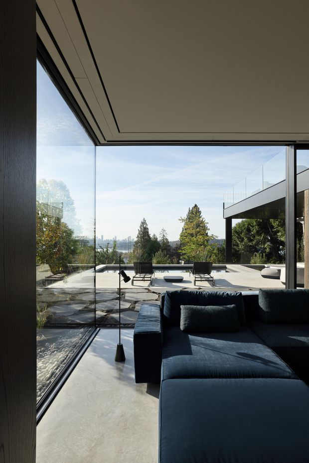 Contemporary living room opening onto a spacious outdoor terrace through floor-to-ceiling glazing. The room features a large dark navy sectional sofa, a slim black floor lamp, smooth grey concrete flooring, and a clean-lined recessed ceiling detail. Outside, a paved terrace with lounge chairs, glass balustrades, and landscaped trees overlooks a distant urban skyline.
