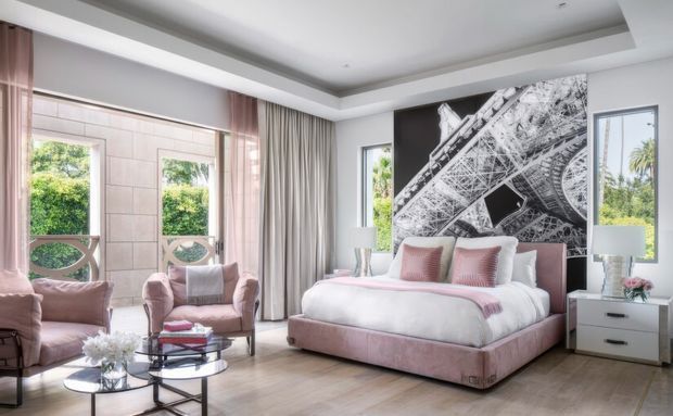 A bright, refined bedroom with a large upholstered bed in blush pink, matching armchairs, layered curtains, pale timber flooring, and a dramatic black-and-white architectural feature mural behind the bed. Large windows bring in greenery and daylight, while mirrored and glass-topped occasional furniture adds a polished feel.