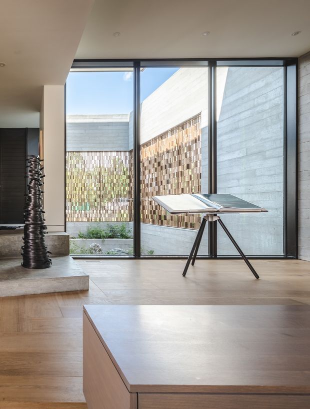 A refined contemporary interior with warm timber flooring and built-in timber joinery, framed by full-height black window glazing. A sculptural dark metal column-like artwork stands on a raised concrete plinth, while a minimalist drafting or display table sits in front of a concrete courtyard wall and patterned privacy screen. The space is bright, carefully detailed, and gallery-like.