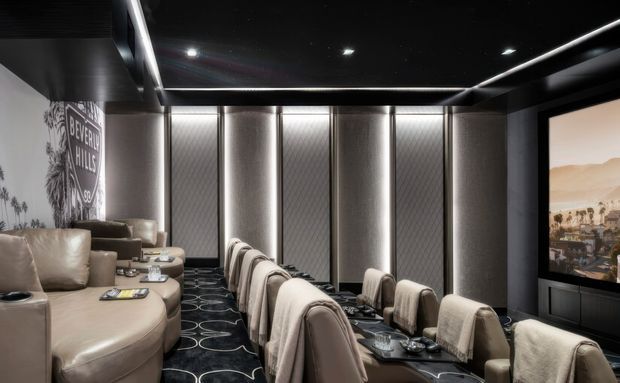 A luxurious private home cinema with tiered rows of plush cream and beige seating, dark acoustic wall panels, integrated vertical lighting, a black ceiling with recessed spotlights and linear LED strips, patterned carpet, and a large projection screen displaying a desert landscape. The room has a dramatic, high-contrast cinematic atmosphere with refined built-in finishes.