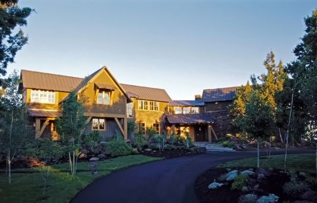 Exterior view of a large, multi-wing timber-clad house set within a landscaped garden. The building has prominent gable roofs, projecting upper-level volumes, covered entry porches, and a dark asphalt driveway curving toward the entrance. Warm late-day light highlights the natural wood tones and surrounding mature trees.