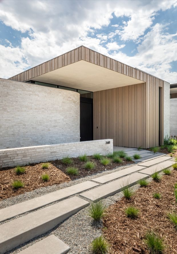 Contemporary residential front entry viewed from the landscaped walkway. The home features a crisp gabled roof form with vertical timber cladding, a broad sheltered entry canopy, pale masonry walls, a dark recessed front door, and minimalist concrete stepping slabs set through gravel and mulched planting.