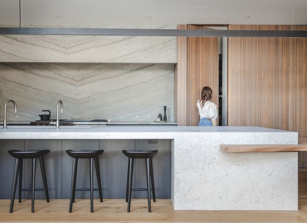 A contemporary kitchen centred on a long stone island with three black bar stools. The island incorporates a sink and tap, while the rear wall features a matching stone splashback and open shelf. Vertical timber wall panelling and cabinetry add warmth, with a concealed doorway and a person providing scale. Pale timber flooring and a slim dark horizontal beam complete the refined, minimalist composition.