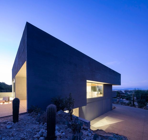 Twilight exterior view of a sharply geometric contemporary house with a dark blue monolithic facade and projecting angular roof or upper volume. Warm interior lighting glows through a large rectangular window and an open side terrace. The home is set on a rocky, drought-tolerant landscape with columnar cacti, and a paved driveway slopes toward a lower-level garage or entry. Distant suburban and mountainous terrain is visible beyond the property.