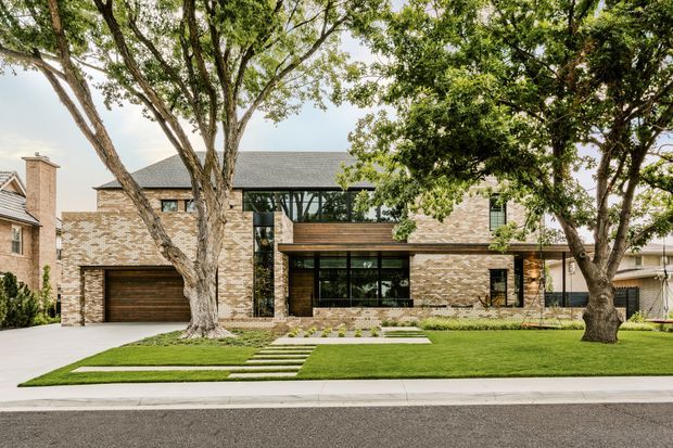 Front exterior view of a large contemporary two-storey residence, featuring pale textured brick masonry, dark tiled pitched roof sections, extensive black-framed glazing, timber garage and entry doors, and a carefully landscaped lawn with concrete stepping paths. Mature street trees partially screen the facade.