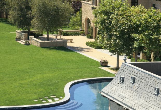 A landscaped residential courtyard or garden viewed from an elevated position, featuring a curving swimming pool with blue water, expansive lawn, clipped trees and shrubs, pale stone paving, low stone retaining walls, outdoor bench seating, and a substantial stone-clad building. A slate-shingle roof structure projects beside the pool in the foreground.