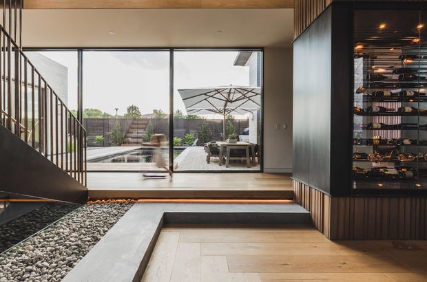 Modern residential interior connecting an entry or living space to a landscaped courtyard and pool through expansive floor-to-ceiling glazing. The room features warm timber flooring and ceiling beams, a dark metal stair with vertical balustrades, a built-in stone pebble feature beside the steps, and a dramatic illuminated glass wine display cabinet with dark timber or metal framing. Outdoor dining and paving are visible beyond the glazing.