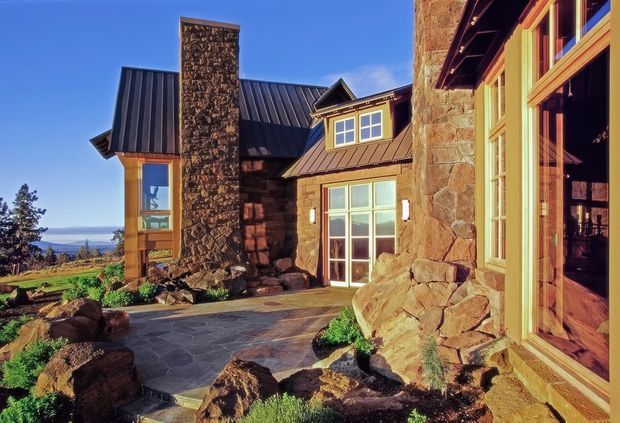 Exterior view of a rustic mountain-style stone and timber residence with multiple dark standing-seam metal roof forms, a tall stone chimney, warm timber-framed glazing, and a flagstone courtyard or entry terrace. Large natural boulders and low landscaping surround the building, with a distant landscape visible beyond.