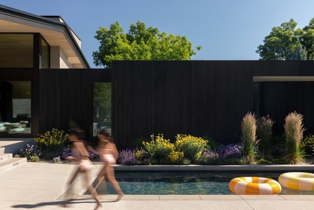 Contemporary residential exterior with a dark timber-clad privacy wall, low horizontal rooflines, expansive glazing, and a narrow swimming pool bordered by structured ornamental planting. The sunlit pool terrace includes blurred figures and bright inflatable pool floats, with mature trees visible beyond the property.