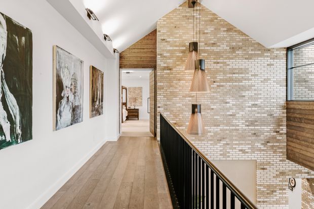 A bright upper-level hallway and stair landing with a pitched ceiling and exposed timber gable feature. The space has wide light timber flooring, white walls with artwork, a black metal stair balustrade, a pale exposed brick feature wall, and three suspended timber pendant lights. A lower stairwell and adjoining rooms are visible beyond the landing.