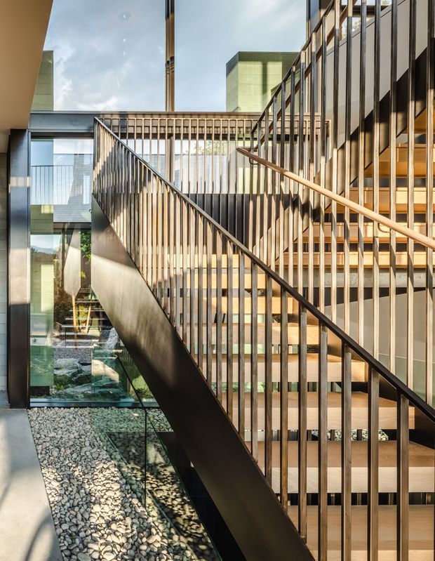 A contemporary multi-level interior centered on a staircase with warm timber treads, dark metal stringers and slender vertical balustrades. Floor-to-ceiling glazing, a glass-lined internal courtyard or lightwell filled with decorative stone, and strong sunlight create a layered, airy architectural space.