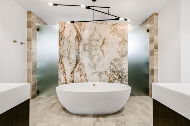 A spacious, symmetrical contemporary bathroom centered on a freestanding white oval bathtub. The bath is backed by a dramatic veined natural-stone feature wall, with frosted-glass shower screens on both sides and matching wall-mounted vanities at the edges. Large-format pale stone-look floor and wall tiles, dark timber-toned vanity fronts, white walls, and slim black geometric ceiling lighting create a refined, high-contrast scheme.