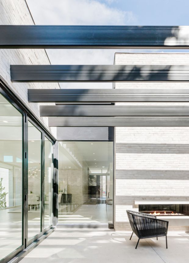 Bright contemporary courtyard or patio adjoining a glazed interior. The space features a white brick wall with a linear recessed fireplace, broad concrete paving, full-height glazed sliding doors, and a series of exposed dark metal pergola beams casting shadows across the terrace. A black woven lounge chair sits near the fireplace, while the interior beyond appears open and minimalist.