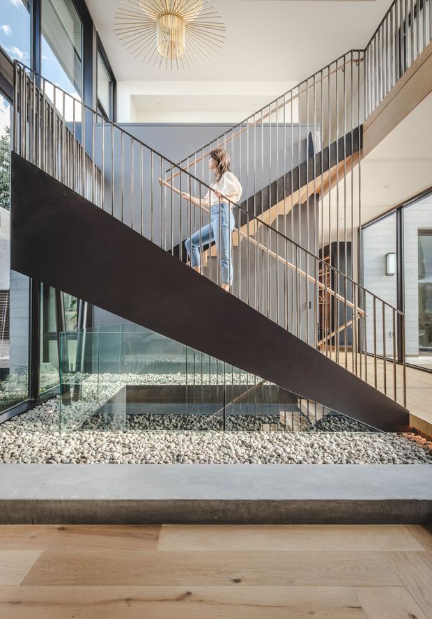 A bright contemporary interior centered on a sculptural open-riser staircase with slim bronze-toned metal balustrades and a dark structural stair stringer. Floor-to-ceiling glazing lines the space, overlooking an indoor pebble-filled water feature or courtyard beneath the stairs. Pale timber flooring, white walls, a large gold sunburst pendant, and an upper-level walkway create a refined, airy architectural setting.